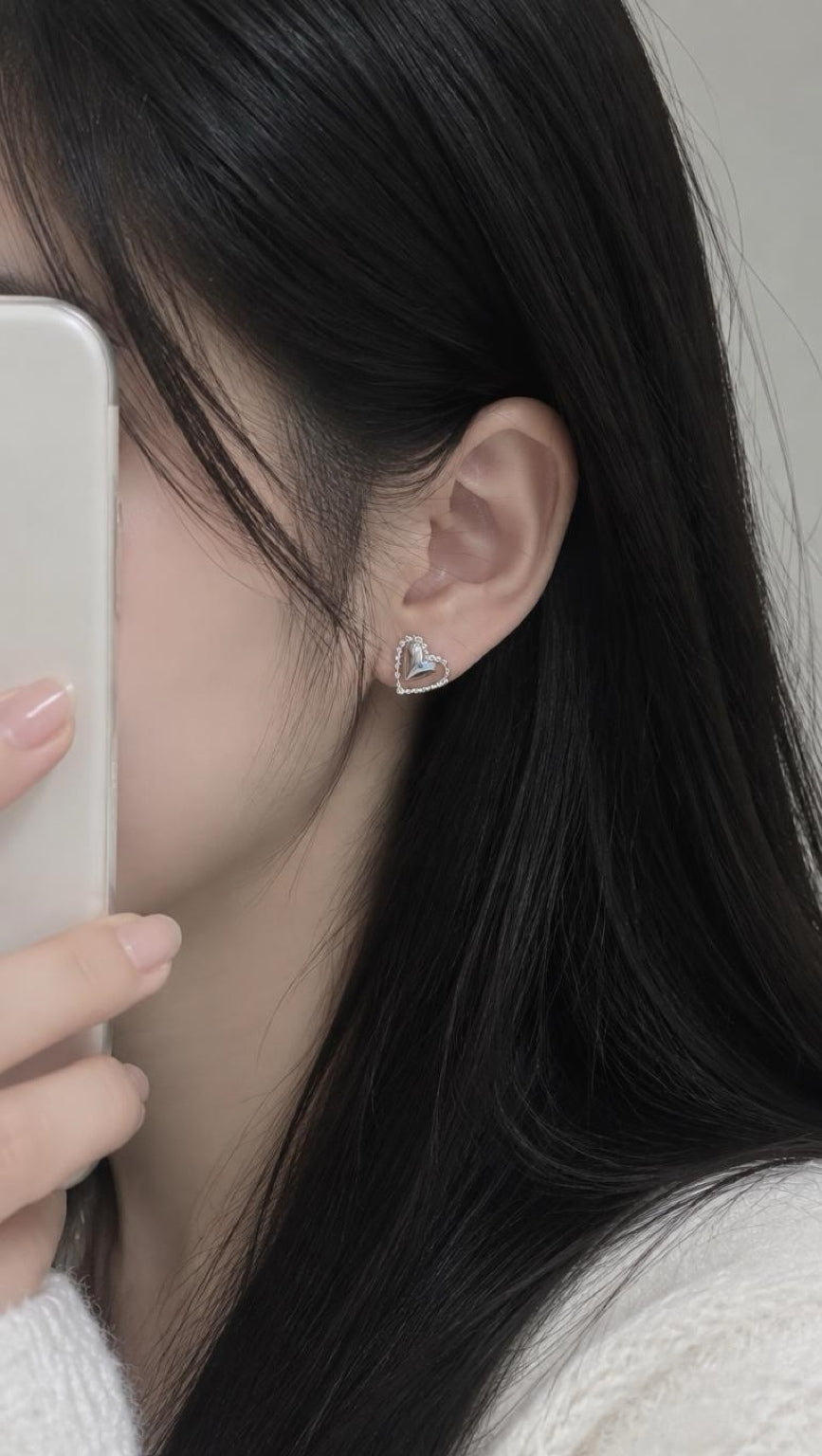 A close-up of a person with long black hair wearing a small silver heart-shaped stud earring, holding a white smartphone to take a mirror selfie.