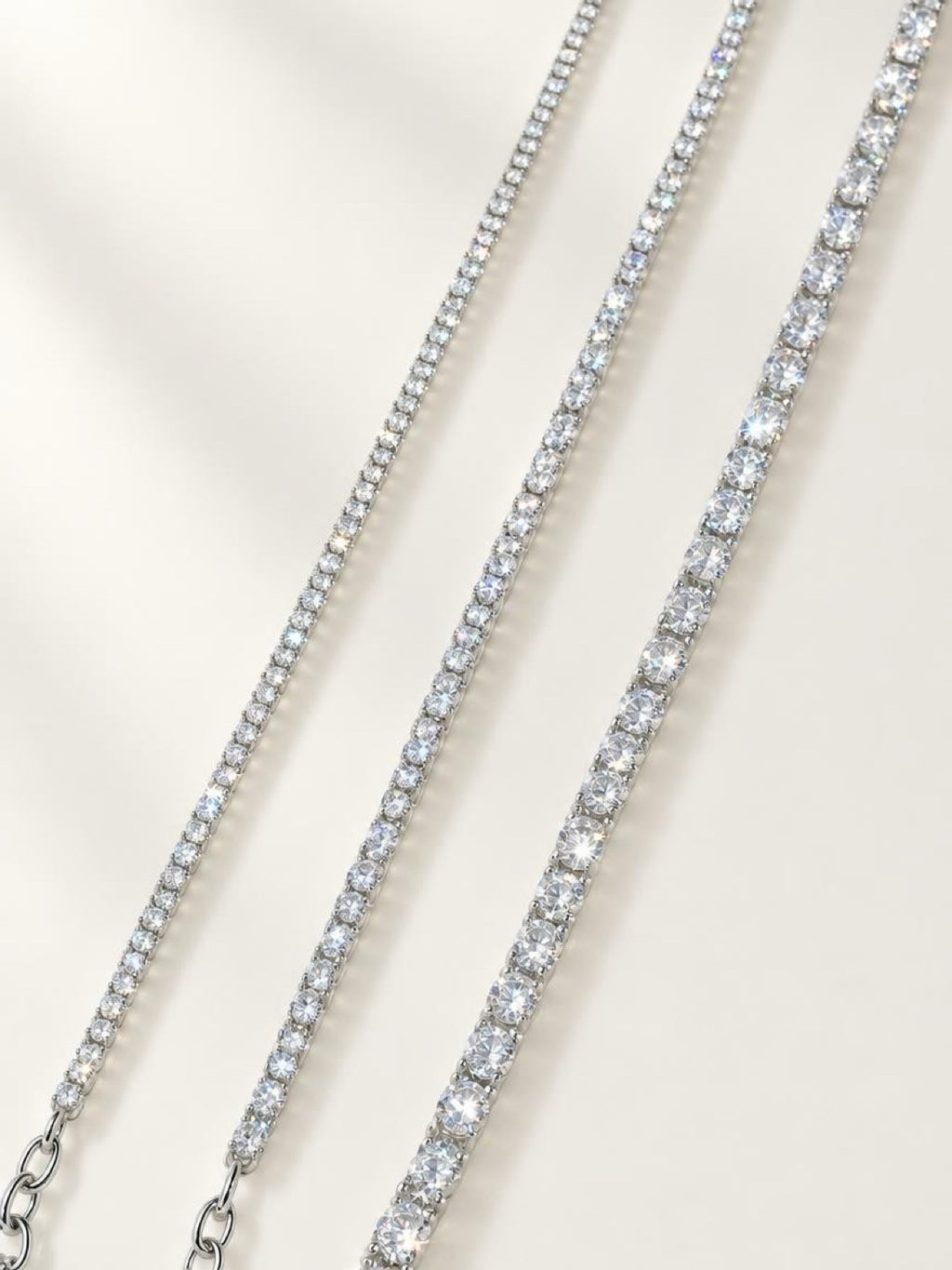 Three tennis-style necklaces with clear stones in silver-toned settings, arranged diagonally on a light cream background.