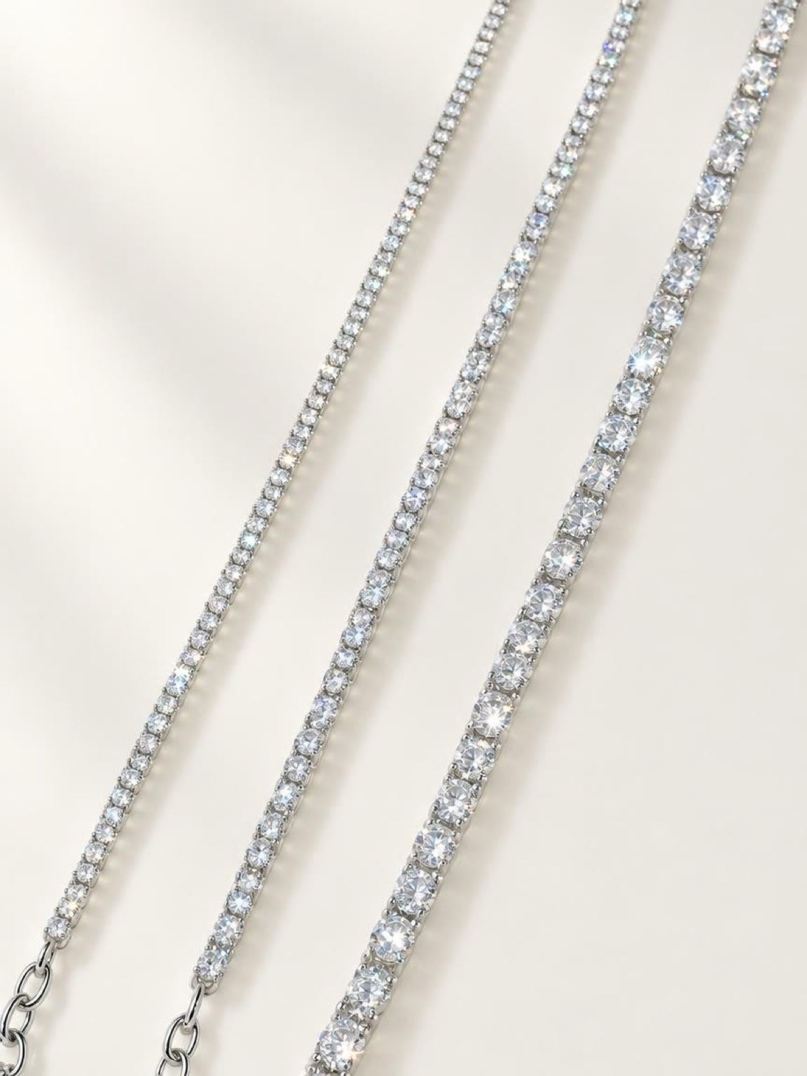 Three tennis-style necklaces with clear stones in silver-toned settings, arranged diagonally on a light cream background.