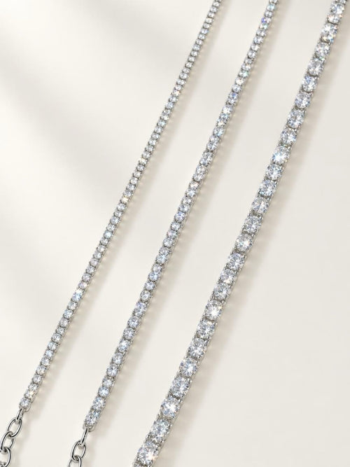 Three tennis-style necklaces with clear stones in silver-toned settings, arranged diagonally on a light cream background.