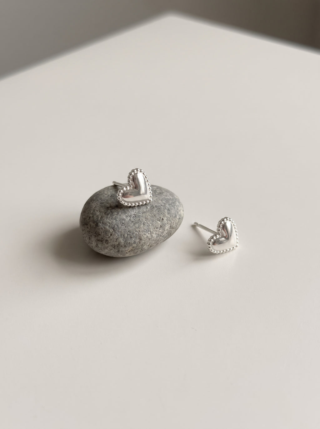 A pair of silver heart stud earrings with beaded edges, one resting on a small gray stone and the other beside it on a light neutral surface.