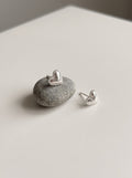 A pair of silver heart stud earrings with beaded edges, one resting on a small gray stone and the other beside it on a light neutral surface.