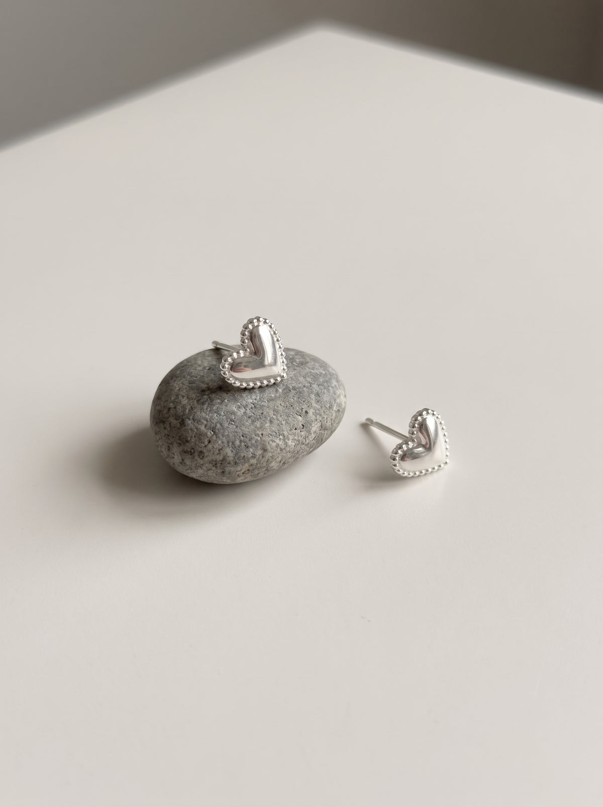 A pair of silver heart stud earrings with beaded edges, one resting on a small gray stone and the other beside it on a light neutral surface.
