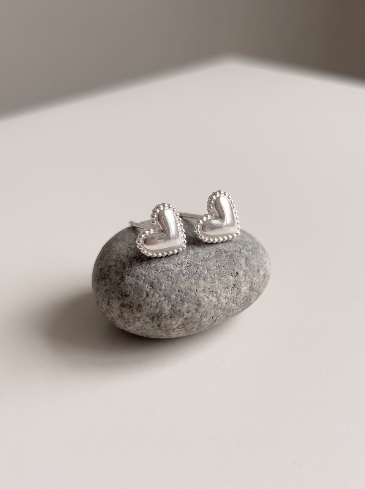 A pair of silver heart-shaped stud earrings with beaded edges, displayed on a smooth gray stone against a neutral light gray background.