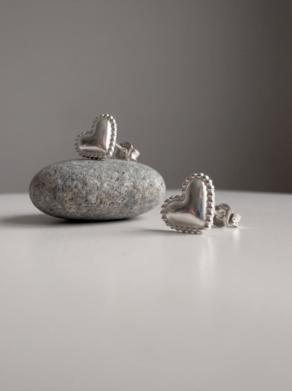 A pair of silver heart-shaped stud earrings displayed on a smooth gray stone and a white surface against a neutral gray background.