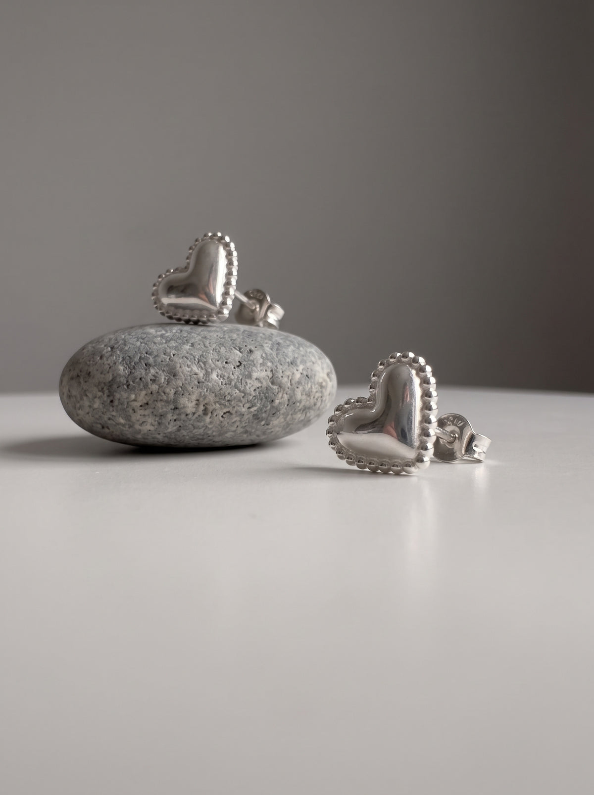 A pair of silver heart-shaped stud earrings displayed on a smooth gray stone and a white surface against a neutral gray background.