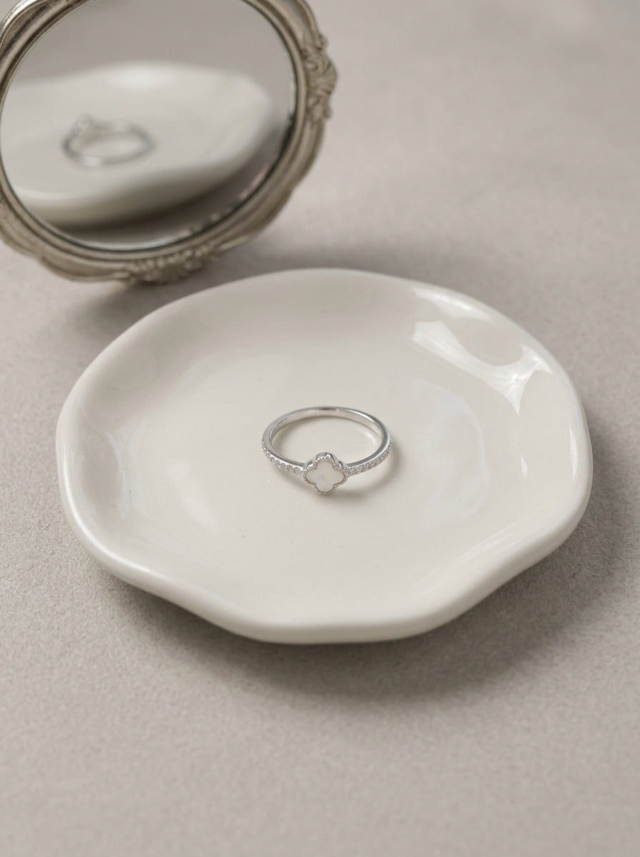 A delicate silver ring with a small clear gemstone sits on a small white ceramic dish, with an ornate silver mirror partially visible in the background on a neutral beige surface.