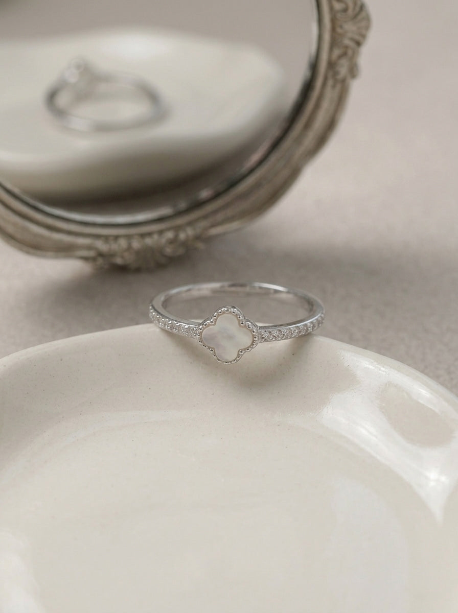 A silver ring with a clover-shaped white mother-of-pearl center stone, resting on a white ceramic dish with an ornate vintage mirror in the background.