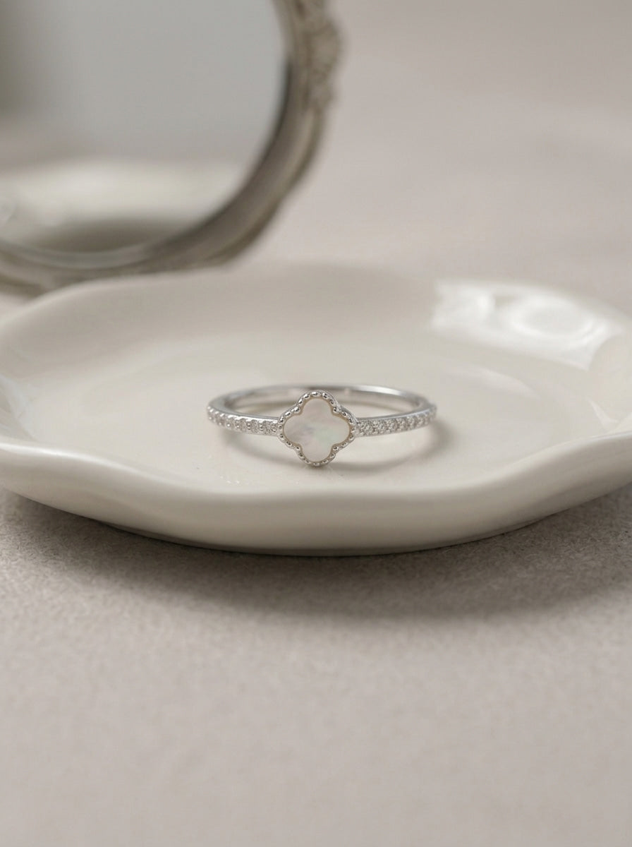 A silver ring with a clover-shaped white stone and small pavé-set stones on the band, displayed on a white ceramic dish.
